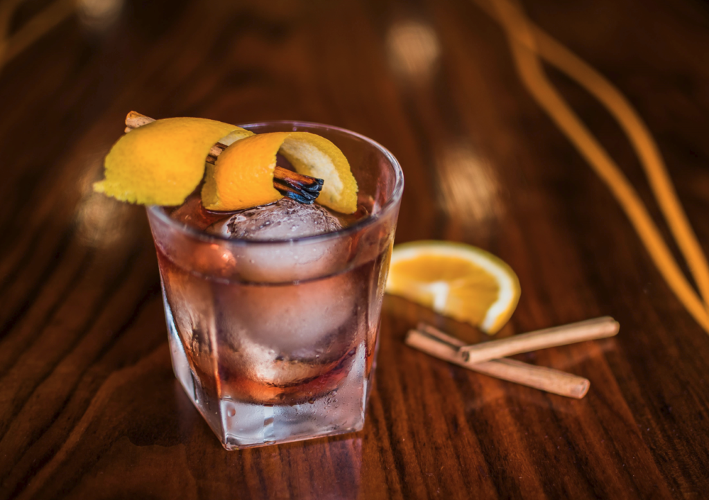 Image of an Old Fashioned drink with an orange twist on top and two cinammon sticks on the side for decorative purposes